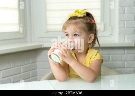 la bambina in una blusa gialla beve il latte da un bicchiere in cucina Foto Stock