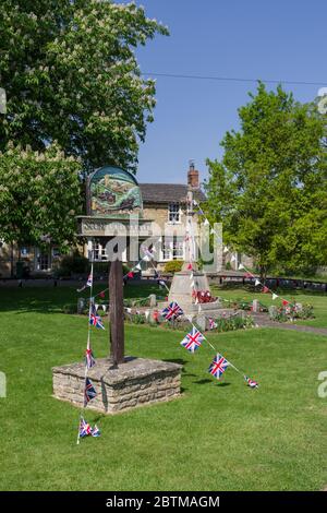 Cartello del villaggio e memoriale di guerra coperto di congestione per celebrare il 75 ° anniversario del VE Day; il villaggio verde, Stoke Goldington, Buckinghamshire, Regno Unito Foto Stock