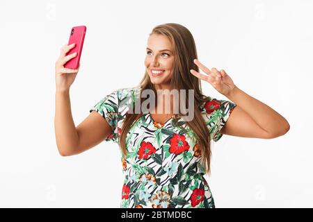 Bella ragazza sorridente in vestito colorato che mostra due dita gesto mentre felicemente scattare foto sul cellulare su sfondo bianco Foto Stock