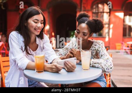 Due belle Signore che usano felicemente il cellulare insieme alle bevande vicino al tavolo mentre si passa il tempo nel cortile del caffè Foto Stock