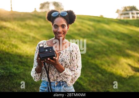 Bella ragazza africana sorridente che tiene in mano la fotocamera polaroid guardare felicemente in macchina fotografica mentre si passa il tempo nel parco Foto Stock