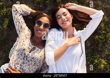 Due belle ragazze sorridenti che si stendono sull'erba mentre trascorrono felicemente del tempo insieme nel parco cittadino Foto Stock