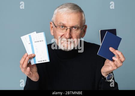 Vecchio uomo sorridente con capelli grigi e barba in occhiali e maglione che tiene biglietti e passaporti in mani mentre si guarda felicemente in macchina fotografica su blu Foto Stock