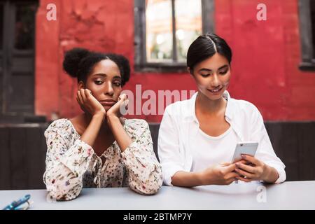 Turbato ragazza africana tristemente guardando da parte, mentre ragazza asiatica vicino felicemente utilizzando il cellulare nel cortile del caffè Foto Stock