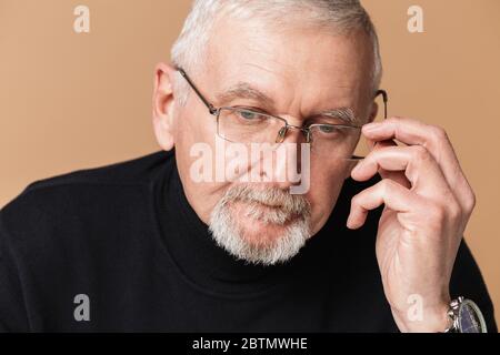 Vecchio uomo pensivo con capelli grigi e barba in occhiali e maglione guardando da parte con cura mentre si passa il tempo su sfondo beige Foto Stock