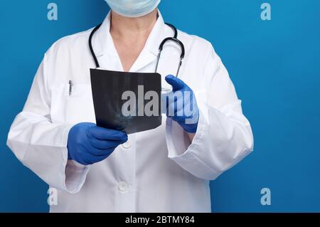 il medico in un camice bianco e i guanti in lattice blu contengono un raggio di raggi x della mano di un uomo e conduce un esame visivo, sfondo bianco Foto Stock