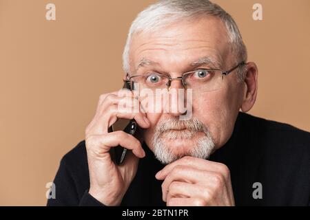 Vecchio uomo pensieroso con capelli grigi e barba negli occhiali e maglione che guarda stupito in macchina fotografica mentre parla sul cellulare su sfondo beige Foto Stock