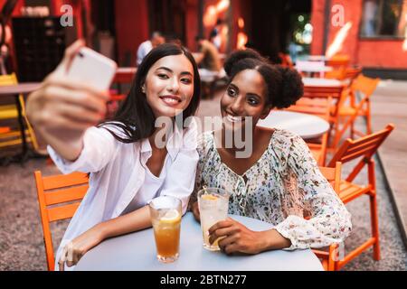 Due giovani Signore seduti a tavola con cocktail che scattano foto felici sul cellulare mentre si passa il tempo insieme in un accogliente cortile del caffè Foto Stock