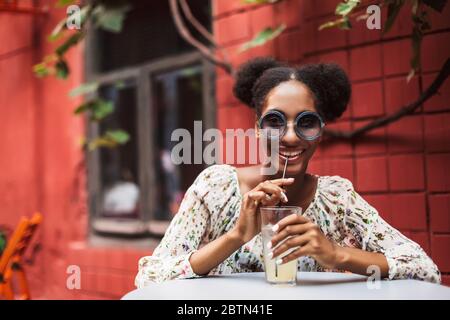 Giovane donna africana in blusa e occhiali da sole che guarda felicemente in macchina fotografica mentre beve un cocktail nell'accogliente cortile del caffè Foto Stock