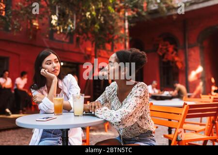 Due belle ragazze sedute a tavolo con cocktail e telefono cellulare che parlano sognatamente mentre si passa il tempo insieme in un accogliente cortile di caffè Foto Stock