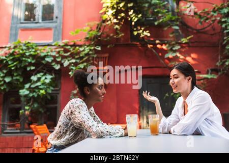 Due belle ragazze che parlano felici con i cocktail mentre trascorrono il tempo nell'accogliente cortile del caffè Foto Stock