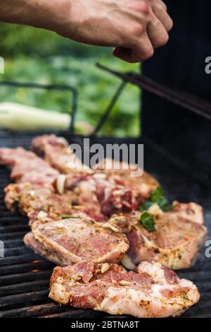 Bistecche di carne sono grigliate su una griglia esterna di strada. L'uomo cucina barbecue e aggiunge spezie. Picnic estivo in natura Foto Stock