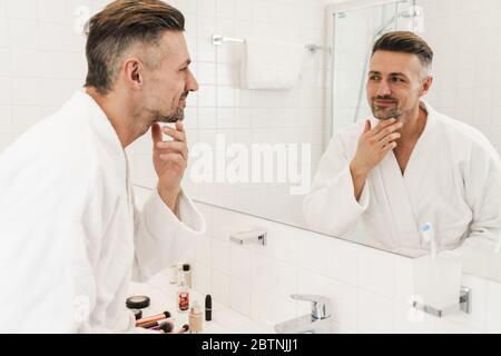 Foto di un uomo caucasico che indossa accappatoi bianchi sorridendo e guardando lo specchio nel bagno Foto Stock