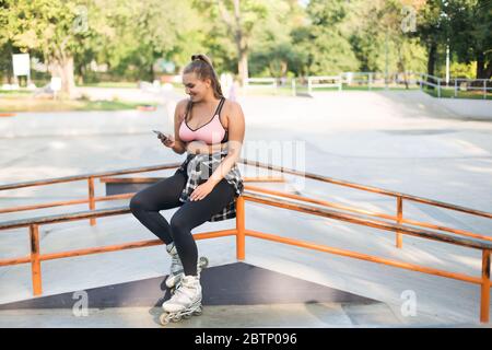 Giovane bella sorridente più taglia donna in rosa sportivo top, leggings e pattini a rotelle appoggiati sulla ringhiera mentre felicemente utilizzando il cellulare in skate Park Foto Stock