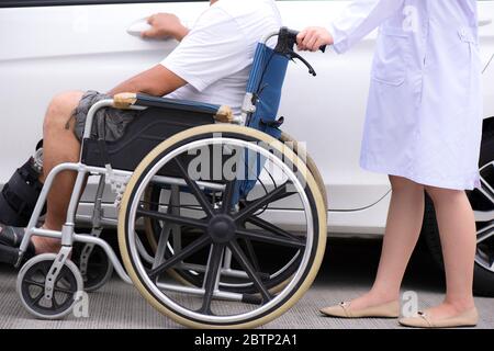 Un uomo anziano seduto su una sedia a rotelle aprire la porta dell'auto sulla strada, con un'infermiera che spinge una sedia a rotelle nella parte posteriore Foto Stock