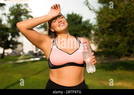 Giovane donna abbastanza sorridente più grande in rosa top sportivo che tiene in mano una bottiglia di acqua pura occhi di chiusura sognanti mentre si passa il tempo nel parco della città Foto Stock