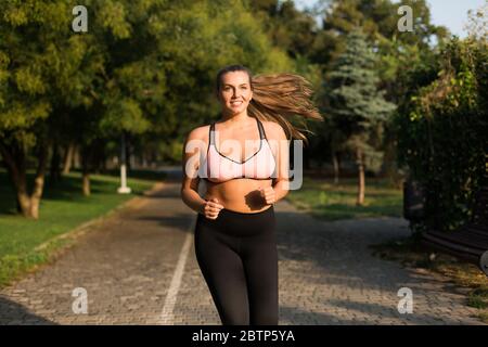 Giovane donna abbastanza sorridente più taglia in top sportivo rosa e leggings felicemente correre nel parco cittadino Foto Stock