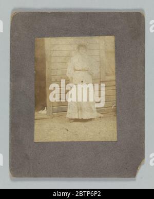 Stampa fotografica di una donna davanti ad un edificio. Una stampa fotografica di una donna non identificata che si trova di fronte ad un edificio con la mano su di lei. Indossa un abito lungo con una cintura in vita e un cofano sulla testa. La stampa è montata su carta nera. Il retro del supporto è vuoto. Foto Stock