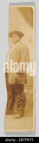 Pezzo di una cartolina fotografica di una donna non identificata in una giacca e pantaloni. Un pezzo di cartolina fotografica con una donna non identificata che indossa una giacca, pantaloni e un ampio cappello a brimmetto. La cartolina è stata strappata sul lato sinistro. La donna si alza verso la fotocamera con la mano sinistra nella tasca della giacca. Foto Stock