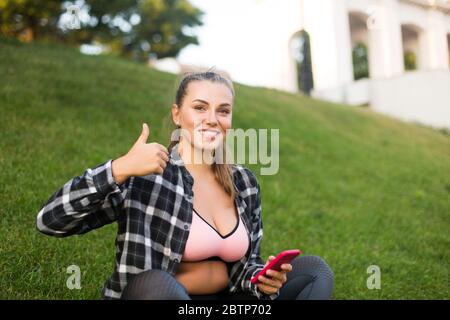 Giovane donna bella più taglia in camicia casual e top sportivo che tiene il cellulare in mano felicemente guardando in macchina fotografica mostrando grande pollice su gesto mentre Foto Stock