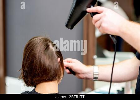 Mani del parrucchiere che asciugano i capelli corti marroni con l'asciugacapelli di soffiaggio in salone di parrucchiere. Foto Stock