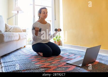 Bella donna spagnola che guarda il corso online su computer portatile, allenarsi con i manubri in soggiorno Foto Stock