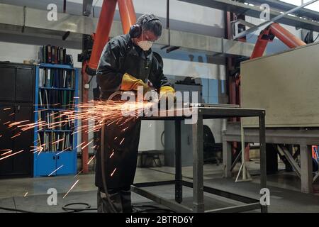 Operatore manuale in maschera e protezioni auricolari in piedi su tavola in metallo alto e taglio di metallo con utensile rotante in officina industriale Foto Stock