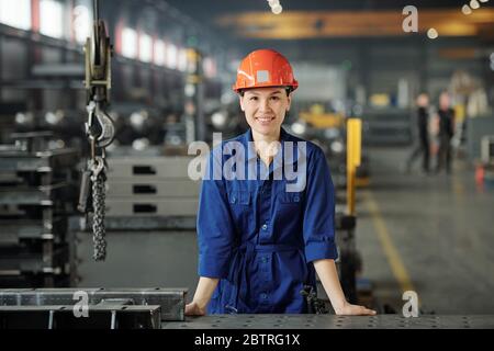 Ritratto di sorridente giovane specialista asiatico della lavorazione dei metalli in tute blu in piedi alla scrivania del negozio industriale Foto Stock