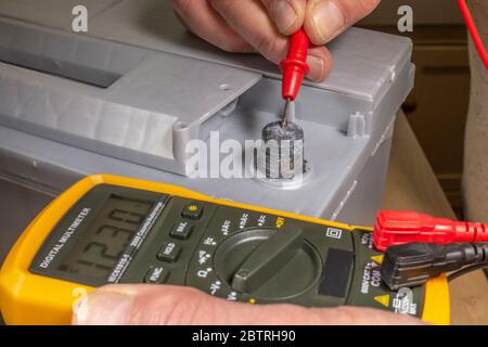 Le mani dell'uomo che prendono la lettura dal multimetro digitale accanto alla batteria del veicolo, per controllare lo stato di carica, con la sonda rossa collegata al morsetto positivo. Foto Stock