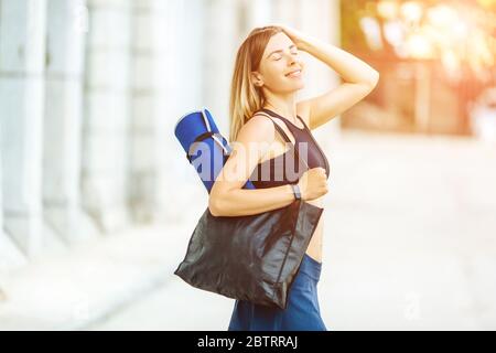 Giovane donna atletica con telefono ad acqua e tappetino sportivo che fa sport all'esterno in tuta sportiva luminosa. Foto Stock