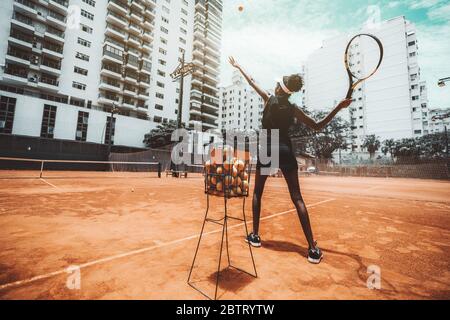 Vista grandangolare da dietro di una giovane donna biraciale sottile che punta un colpo prima di colpire una palla da tennis durante un allenamento sportivo o il riscaldamento su un esterno Foto Stock