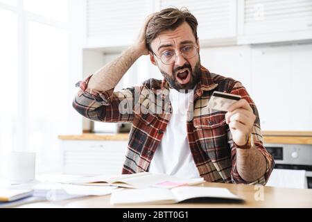 Ritratto di un giovane uomo scioccato che indossa occhiali che tengono la carta di credito mentre lavora a casa Foto Stock