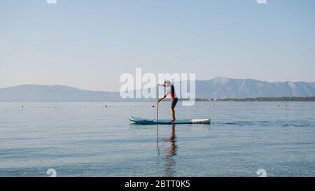 Giovane uomo in abbigliamento casual che si addlea sulla tavola sup su calme acque del mare del mattino. Foto Stock