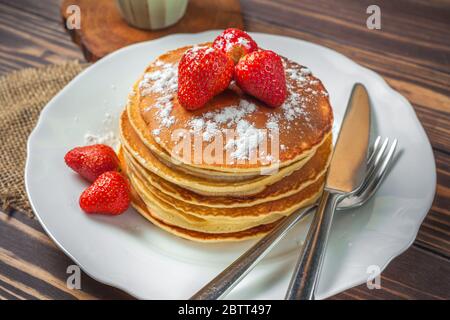 Pancake con fragole, zucchero a velo e una tazza di cappuccino per la colazione su sfondo di legno in stile rustico. Foto Stock