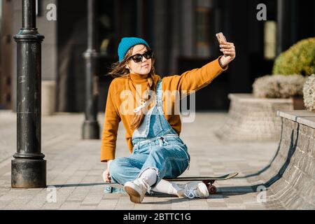 Ragazza seduta sullo skateboard e utilizzare il telefono cellulare. Outdoor, stile di vita urbano. Ragazza skater carina seduta su skate board controllo smartphone ascolto Foto Stock