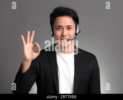 Sorridente giovane uomo d'affari asiatico che parla con le cuffie e mostra il segno ok Foto Stock