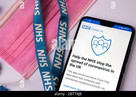 Schermata iniziale dell'applicazione di tracciamento dei contratti NHS COVID-19 visualizzata sul dispositivo mobile, accanto al cordino blu NHS e alla maschera rosa. Messa a fuoco selettiva. Foto Stock