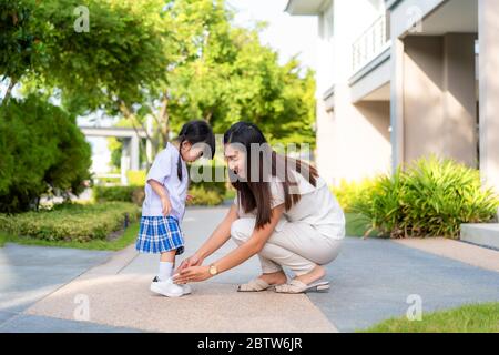 Madre asiatica che aiuta sua figlia a mettere scarpe su o decollo al parco all'aperto pronti a uscire insieme o tornare a casa da scuola in felice f Foto Stock
