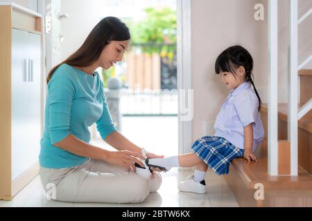 Madre asiatica che aiuta sua figlia a mettere scarpe su o decollo a casa per prepararsi a uscire insieme o tornare a casa da scuola in famiglia felice wi Foto Stock