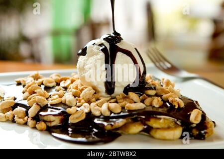 Crepe al cioccolato e gelato alla vaniglia, fette di banana e guarnizioni di arachidi. Sciroppo gocciolante dall'alto. Gustoso dessert gourmet dolce, cucina francese Foto Stock