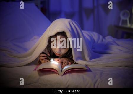 Bambino asiatico ragazza che legge un libro con torcia sotto la coperta sul letto in una camera da letto scura di notte prima di dormire, bambini confortevoli a casa conc Foto Stock