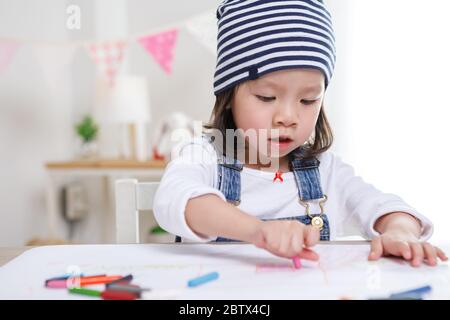 Bambina asiatica seduta a tavola in camera, ragazza preschooler che si occupa di carta con penne colorate il giorno di sole, asilo o cura di giorno. Educativo Foto Stock