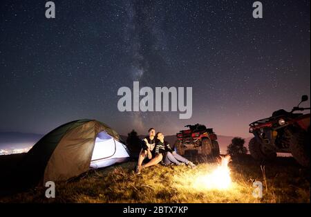 Uomo e donna seduti insieme vicino a tenda turistica, fuoco di fuoco, due quad atv moto sulla cima della montagna, coppia romantica godendo di una bella vista del cielo notturno pieno di stelle e Via Lattea Foto Stock