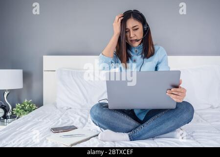 donna di affari stressata indossa la cuffia videochiamata di conferenza sul computer portatile su un letto, lavoro da casa concetto Foto Stock