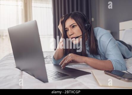 donna di affari stressata indossa la cuffia videochiamata di conferenza sul computer portatile su un letto, lavoro da casa concetto Foto Stock