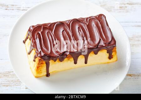 Cheesecake senza flourless di formaggio ucraino cottage con uvetta e glassa di cioccolato fondente sulla parte superiore servita su un piedistallo di torta bianca su sfondo di legno bianco Foto Stock