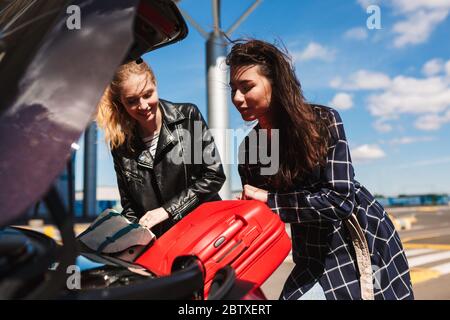 Belle ragazze gioiosamente pieghevole valigie insieme nel bagagliaio di auto vicino aeroporto Foto Stock