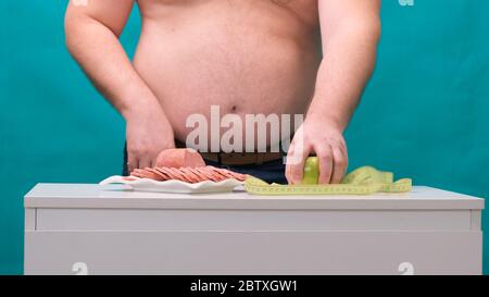 l'uomo con una pancia grassa sceglie tra salsiccia e mela verde. Il concetto di dieta e di volontà. Foto Stock