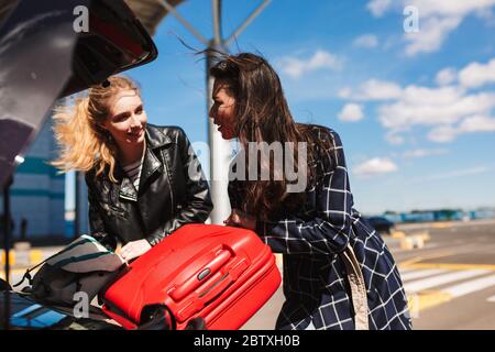 Ragazze graziose che si guardano sognatamente mentre si piegano le valigie nel bagagliaio di auto nera vicino all'aeroporto Foto Stock