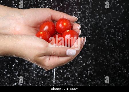 Donna mano che tiene il pomodoro di ciliegia rosso sotto le gocce d'acqua in uno sfondo nero. Cibo vegetariano. Stile di vita sano. Foto Stock